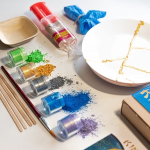 May include: A set of six small jars of colorful powder, a bottle of glue, two plates, and a wooden box with the text "Kintsugi" on it. The powder is for creating a kintsugi effect on the plates.