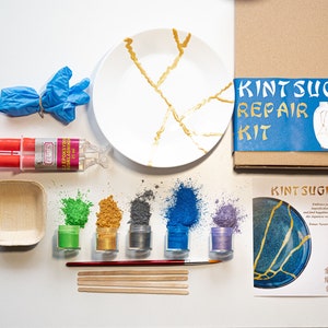 May include: Kintsugi repair kit with a white plate with gold cracks, a tube of epoxy resin, five small jars of metallic powder in green, gold, gray, blue, and purple, a wooden bowl, four wooden sticks, a paintbrush, and instructions. The kit is labeled "Kintsugi Repair Kit".