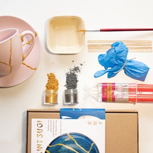 Puede incluir: Un kit de Kintsugi con una taza de té y un platillo rosas con grietas doradas, un pequeño cuenco de madera, dos frascos de polvo dorado y gris, una botella de resina epoxi, un par de guantes azules, dos palitos de madera, un pincel y una caja de cartón con el texto "Kintsugi" y un plato azul y dorado.