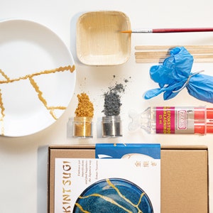 May include: Kintsugi kit featuring a white ceramic plate with gold cracks, two small jars of gold and grey powder, a small wooden bowl, a bottle of epoxy resin, a red and white epoxy resin dispenser, a pair of blue gloves, wooden sticks, a paintbrush, and a brown box with the text "Kintsugi" and a picture of a blue plate with gold cracks.