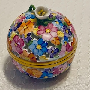 May include: A round, floral-patterned trinket box with a lid. The box is white with colorful flowers in shades of pink, blue, orange, and purple. A small white flower with yellow stamens sits atop the lid. The box has a yellow rim.