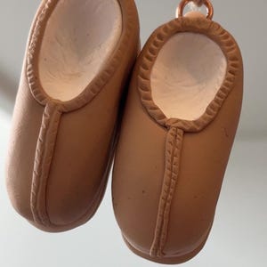 Puede incluir: Dos zapatos de bebé de cerámica marrón colgando de una cuerda. Los zapatos tienen un interior blanco y están conectados a anillos de color cobre. Los zapatos son de color marrón claro y tienen un diseño cosido en el centro.