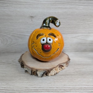 Könnte beinhalten: Ein Keramik-Kürbis mit einem lächelnden Gesicht, schwarzen Punkten, weißen Augen und einer roten Nase. Der Kürbis ist orange mit einem grünen Stiel und sitzt auf einer Holzscheibe. Ein festlicher Dekorationsartikel.
