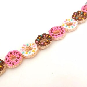 May include: A beaded bracelet with donut charms. The donuts are made of beads in pink, white, and brown with colorful sprinkles.