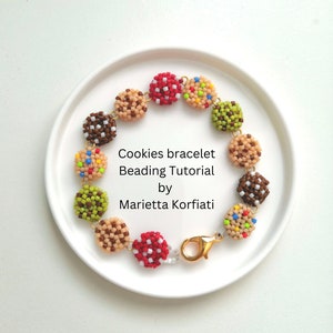 May include: A beaded bracelet with cookie charms in various colours, including brown, red, green, and beige. The bracelet has a gold clasp and is displayed on a white plate. The text "Cookies bracelet Beading Tutorial by Marietta Korfiati" is written below the bracelet.
