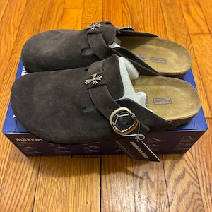 May include: Dark brown suede clogs with silver-tone metal detailing. The clogs feature a buckle fastening and are presented on a blue box. The footbed is tan. New clogs with tags.