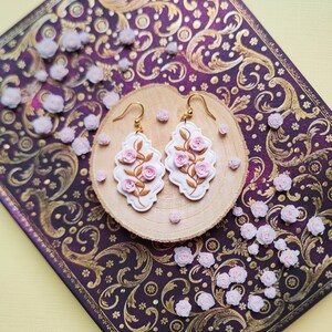 Könnte beinhalten: Weiß-goldene Ohrringe mit rosa Rosenakzenten. Die Ohrringe liegen auf einer Holzscheibe, umgeben von kleinen rosa Rosenverzierungen. Der Hintergrund zeigt eine dekorative Oberfläche mit einem lila-goldenen Muster.