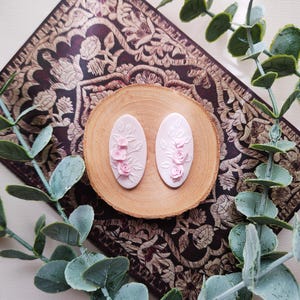 Könnte beinhalten: Zwei ovale, hellrosa Ohrringe mit floralen Rosendesigns, präsentiert auf einer Holzscheibe. Die Ohrringe sind vor einem dunklen, gemusterten Hintergrund mit grünen Blättern platziert.