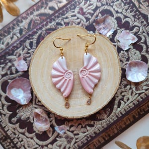 May include: A pair of pink seashell-shaped earrings with gold-colored hooks and small dangling charms. The earrings are displayed on a wooden slice, surrounded by decorative elements. The earrings have a pearlescent finish.