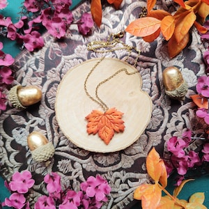 May include: A fall-themed necklace with an orange maple leaf pendant on a gold chain, displayed on a wooden slice. The image includes gold-tipped acorns and pink flowers, with orange and yellow leaves, creating a seasonal aesthetic.