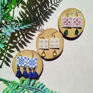 May include: Three pairs of earrings with tile-inspired designs. The first pair features blue and white tiles with gold accents and blue tassels. The second pair features white tiles with gold accents and yellow teardrop-shaped stones. The third pair features pink and white tiles with gold accents and green teardrop-shaped stones.