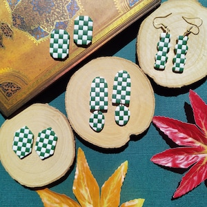 Könnte beinhalten: Vier Paar grüne und weiße karierte Ohrringe aus Polymer Clay. Die Ohrringe werden auf Holzrundlingen und einem Buch mit goldener Oberfläche präsentiert.