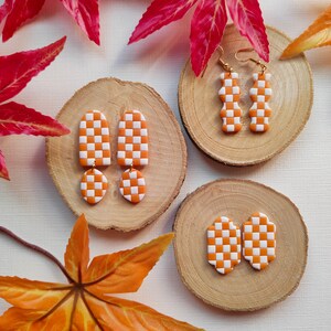 Könnte beinhalten: Drei Paar orange-weiße karierte Ohrringe aus Polymer Clay. Die Ohrringe werden auf drei Holzrundlingen präsentiert. Die Ohrringe haben die Form eines Rechtecks, eines Bogens und einer Träne.