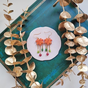 May include: Orange floral earrings with gold-colored hooks and dangling chains. The earrings feature green, pink, and yellow beads. The earrings are displayed on a white, circular surface against a teal background with gold accents and gold-colored leaves.