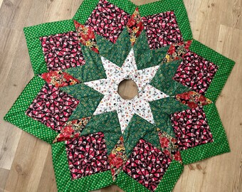 May include: A green, red, and white star-shaped Christmas tree skirt with a gold trim. The skirt is made of fabric with a variety of patterns, including gnomes, snowflakes, and swirls.