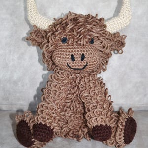 May include: A brown crocheted stuffed animal resembling a Highland cow with white horns and brown hooves. The cow has a smiling face with black eyes and a black nose.