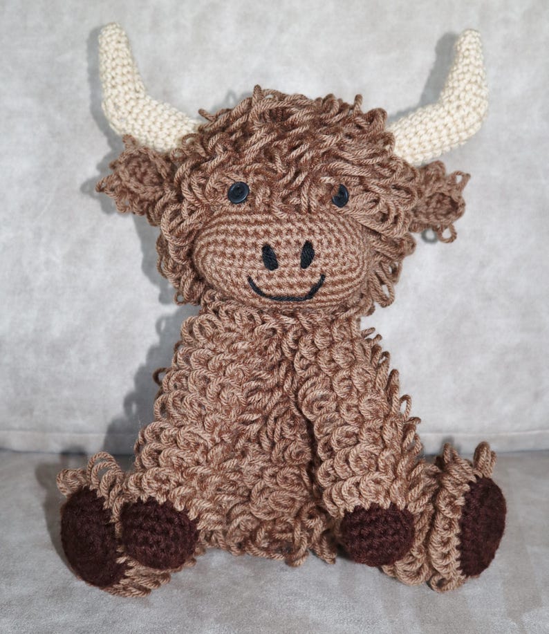 May include: A brown crocheted stuffed animal resembling a Highland cow with white horns and brown hooves. The cow has a smiling face with black eyes and a black nose.