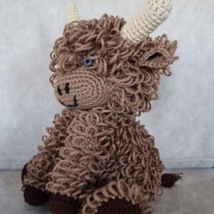 May include: A brown crocheted stuffed animal resembling a Highland cow with white horns and blue eyes. The animal is sitting with its legs tucked under its body.