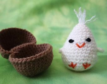 May include: A handmade crochet chick with a brown eggshell. The chick is white with black eyes, an orange beak, and orange feet. The eggshell is brown and open, revealing the inside. The items are on a green background.