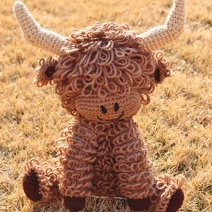 May include: A brown crocheted stuffed animal resembling a Highland cow with long, curly fur. The toy has a white crocheted nose and horns, and black embroidered eyes and a mouth.