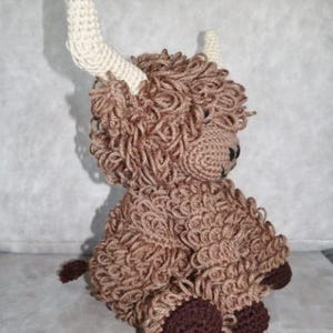 May include: A brown crocheted stuffed animal resembling a Highland cow with white horns. The animal is sitting with its legs tucked under its body and has brown hooves.