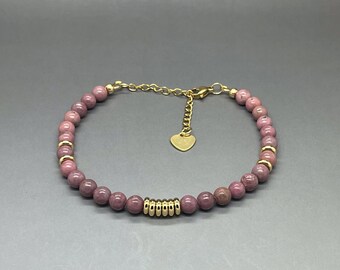 Puede incluir: Un brazalete de cuentas rosadas y doradas con un dije en forma de corazón. El brazalete está hecho con pequeñas cuentas redondas y una cadena dorada.