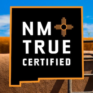 May include: A black and orange sign with the text "NM TRUE CERTIFIED" and a stylized symbol of the sun with rays. The sign is in front of a brown adobe wall and a wooden ladder.
