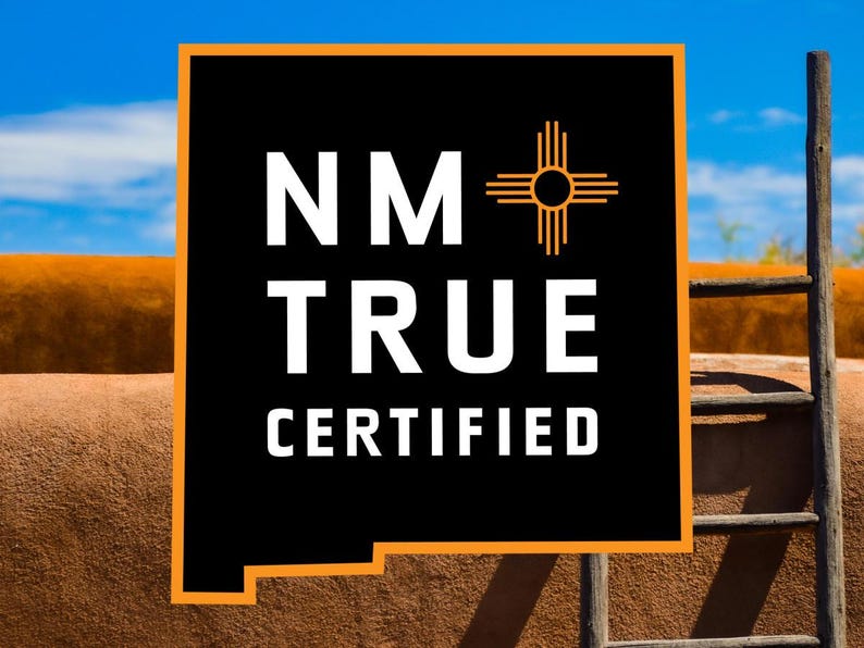 May include: A black and orange sign with the text "NM TRUE CERTIFIED" and a stylized symbol of the sun with rays. The sign is in front of a brown adobe wall and a wooden ladder.
