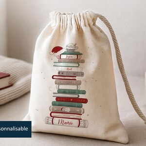 Può includere: Un sacco beige con coulisse con un design ad albero di Natale fatto di libri. I libri sono rossi, verdi e bianchi, con le parole "Joyeux", "Noel" e "Mamie". Un cappello di Babbo Natale e una tazza di caffè sono in cima.