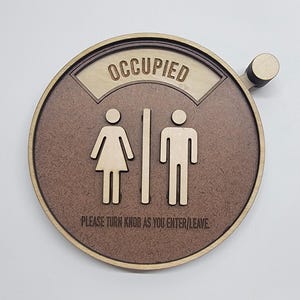 May include: A round wooden sign with a brown background and a white border. The sign says "Occupied" in a white banner at the top.  Below the banner are two stick figures, one female and one male, separated by a vertical line. The sign also says "Please turn knob as you enter/leave".