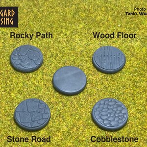 May include: Five gray, circular miniature bases with textured surfaces, including "Rocky Path," "Wood Floor," "Stone Road," and "Cobblestone." The bases are displayed on a green surface, with the "Asgard Rising" logo in the top left corner.