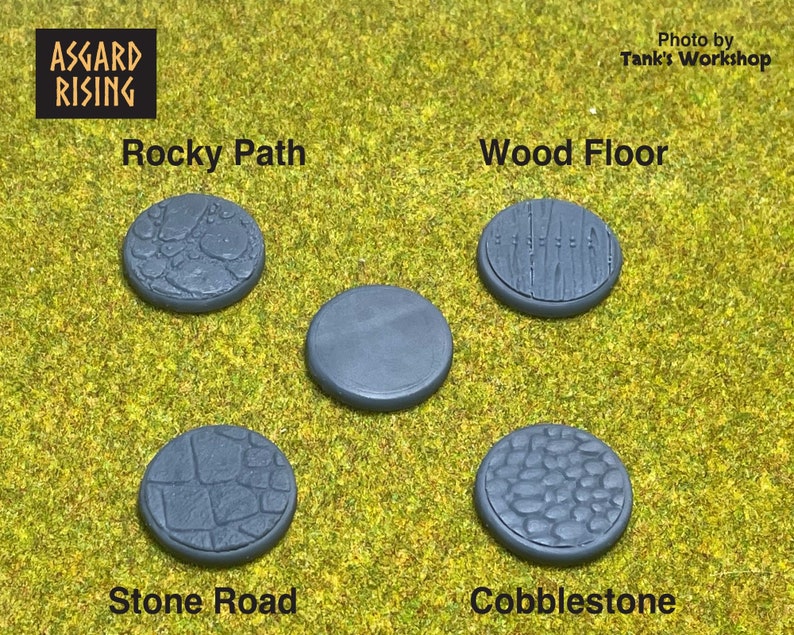 May include: Five gray, circular miniature bases with textured surfaces, including "Rocky Path," "Wood Floor," "Stone Road," and "Cobblestone." The bases are displayed on a green surface, with the "Asgard Rising" logo in the top left corner.