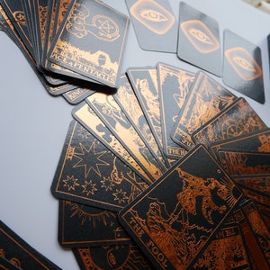 Może przedstawiać: Talia kart tarota z czarnymi kartami i miedzianymi ilustracjami. Karty są rozłożone, prezentując r&oacute;żne obrazy i tekst, w tym "Ace + Pentacles" i "The Fool". Widoczne jest r&oacute;wnież czarne pudełko z napisem "Forever Tarot".