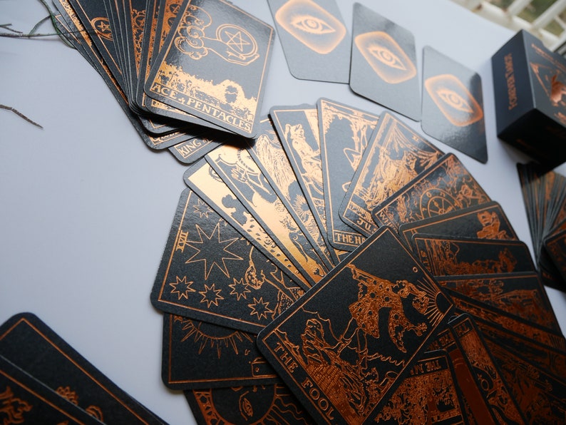 Może przedstawiać: Talia kart tarota z czarnymi kartami i miedzianymi ilustracjami. Karty są rozłożone, prezentując r&oacute;żne obrazy i tekst, w tym "Ace + Pentacles" i "The Fool". Widoczne jest r&oacute;wnież czarne pudełko z napisem "Forever Tarot".