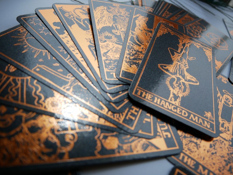 Może przedstawiać: Zestaw kart tarota z czarnym tłem i miedzianymi ilustracjami. Karty zawierają szczeg&oacute;łowe ilustracje i tekst "THE HANGED MAN" na jednej karcie. Karty są rozłożone wachlarzem, prezentując skomplikowane wzory.