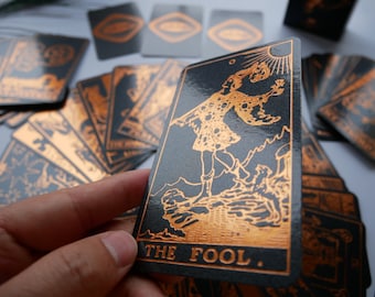 May include: A deck of tarot cards with a black background and copper-colored illustrations. The card in focus features an image of a figure with the text "THE FOOL." Other cards are visible in the background, with similar designs.