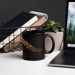 May include: Black ceramic mug with a gold circular design. The mug is on a white surface with a laptop, a succulent plant in a beige pot, and a wire basket containing notebooks.