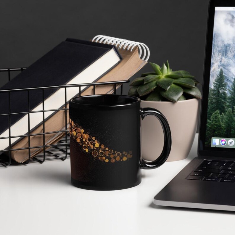 May include: Black ceramic mug with a gold circular design. The mug is on a white surface with a laptop, a succulent plant in a beige pot, and a wire basket containing notebooks.