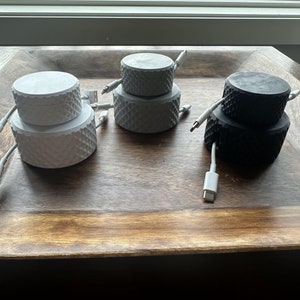 May include: Three cable organizers, one white, one black, and one gray, are stacked on top of each other. The organizers are made of a textured material and have a round shape. The cables are white and are plugged into the organizers.