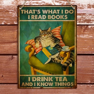 May include: A vintage-style metal sign featuring a cat sitting in a chair, reading a book and drinking tea. The sign reads "That's what I do I read books I drink tea and I know things."