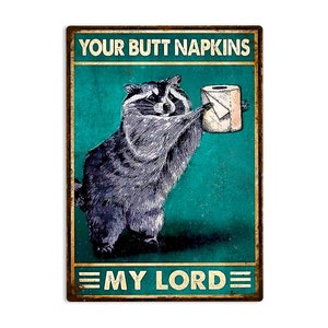 May include: A rectangular metal sign with a distressed finish. It features a raccoon holding a roll of toilet paper, with the text "YOUR BUTT NAPKINS" above and "MY LORD" below. The background is teal.