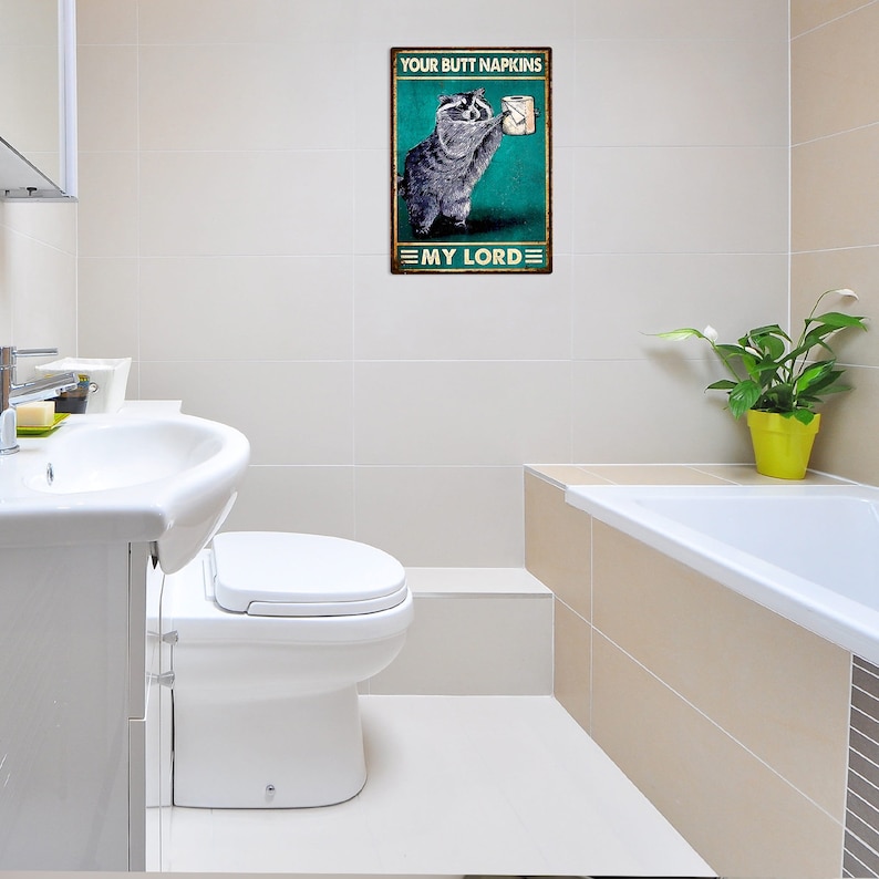 May include: A rectangular metal sign featuring a raccoon holding toilet paper. The sign has a green background and the text "YOUR BUTT NAPKINS" above the raccoon and "MY LORD" below. The sign is in a bathroom.