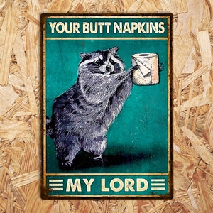 May include: A rectangular metal sign featuring a raccoon offering a toilet roll. The sign reads "YOUR BUTT NAPKINS" above and "MY LORD" below. The background is teal, with a distressed, vintage aesthetic. The sign is designed to look old.