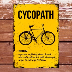 May include: A yellow metal sign with a distressed finish. The sign has black text that reads "CYCOPATH" and a definition of the word. The definition reads "NOUN- a person suffering from chronic bike riding disorder with abnormal urges to ride and feel free." The sign also features a black silhouette of a bicycle.