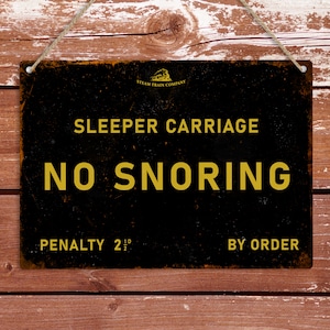 May include: A black metal sign with a yellow border and text that reads "Sleeper Carriage No Snoring Penalty 2 1/2 10 By Order". The sign is hanging from a string on a wooden background.