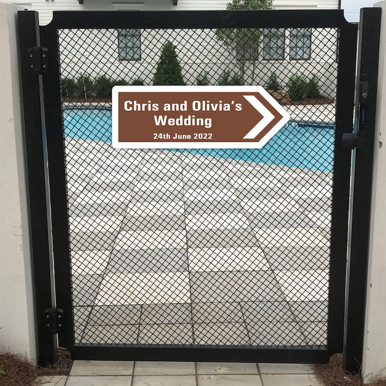 Puede incluir: Un cartel marr&oacute;n y blanco con una flecha que apunta hacia la derecha. El cartel dice "Chris and Olivia's Wedding 24th June 2022". El cartel est&aacute; sujeto a una puerta de metal negro.
