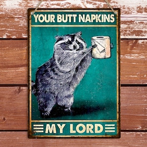May include: Metal sign with a raccoon holding a roll of toilet paper. The sign has the text "YOUR BUTT NAPKINS" at the top and "MY LORD" at the bottom. The background is teal with a distressed look, and the border is gold.