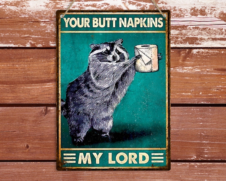 May include: A rectangular metal sign featuring a raccoon offering a toilet roll. The sign has the text "YOUR BUTT NAPKINS" and "MY LORD". The background is teal with a distressed effect, and the border is gold, with a string for hanging.