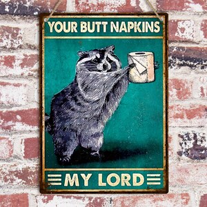 May include: A rectangular metal sign featuring a raccoon holding a toilet roll. The sign reads "YOUR BUTT NAPKINS" at the top and "MY LORD" at the bottom. The background is teal with a distressed effect, and the sign is framed.