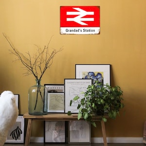 May include: A red and white vintage metal sign with the text "Grandad's Station" and a railway symbol. The sign is hanging on a yellow wall with a vase of branches and framed artwork on a wooden shelf below.